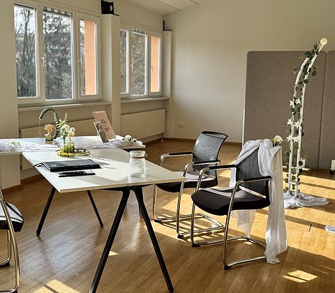 Heller Raum mit zwei Stühlen, weiß dekoriert, einem Tisch mit Ordner und Glas, Fenster und Blumendekorationen.