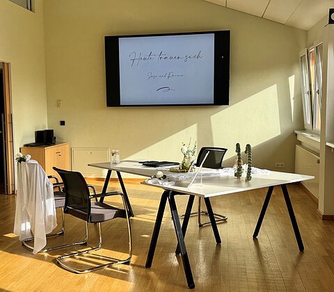 Büro mit zwei Stühlen, einem Tisch mit Dekoration und Bildschirm mit Text „Heute trauen sich“