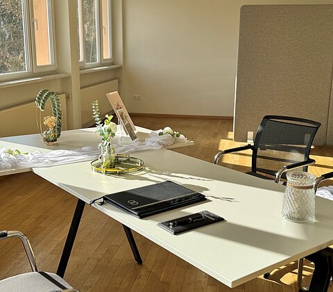 Bürotisch mit Dekoration, Ordner und Stühlen in einem hellen Raum mit Holzfußboden und großen Fenstern.