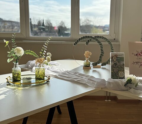 Weißer Tisch mit grünen Glasvasen, weißen Blumen, Dekorationen und einer Karte mit Text zur Hochzeit vor Fenstern.