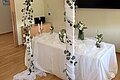 Tisch mit weißem Tuch, dekoriert mit weißen Blumen, Kerzen und mit Stoff umwickelten Ständern mit grünen Blättern.