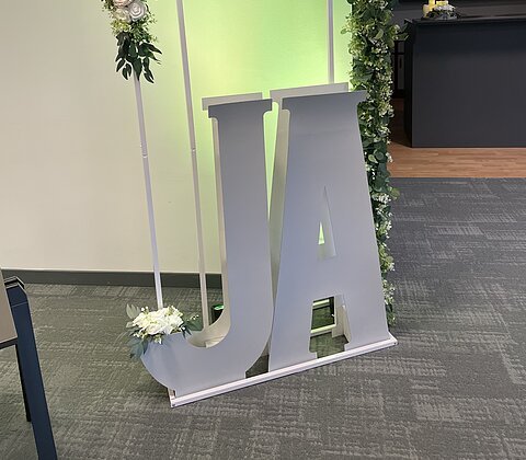 Große weiße Buchstaben „J“ und „A“ mit Blumenbogen aus weißen Rosen und grünem Laub dahinter.