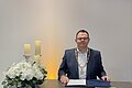 Mann im Anzug mit Amtskette sitzt an Tisch mit Dokumenten, Kerzen und Blumenarrangement vor weißer Wand.