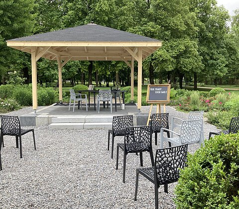 Freiluft-Pavillon mit Stühlen und Tischen auf Kiesplatz, umgeben von Büschen und Bäumen, Tafel mit Text „Ihr traut euch was!“