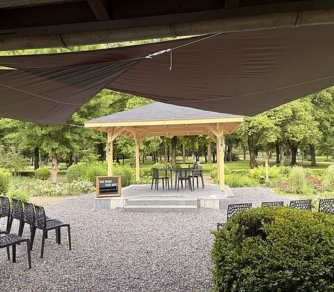 Freiluftbereich mit Holzpavillon, Stühlen und Tafel auf Kiesplatz, umgeben von Bäumen und Pflanzen.