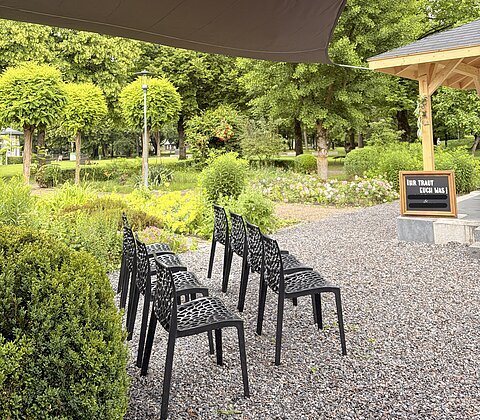 Schwarze Stühle unter Sonnensegel auf Kiesweg neben Pavillon und grüner Parklandschaft mit Büschen und Bäumen.