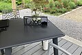 Blick von überdachtem Sitzplatz mit Tisch und Stühlen auf Garten mit Blumenbeeten und Bäumen im Hintergrund.