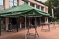 Grüner Sonnenschirm mit Schriftzug vor einem Gebäude mit mehreren Tischen auf gepflastertem Platz bei bewölktem Himmel.
