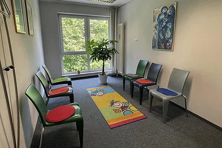 Wartezimmer mit sechs Stühlen, buntem Teppich, Pflanze vor Fenster und abstraktem Bild an der Wand.