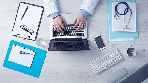 Arzt tippt auf Laptop, umgeben von Stethoskop, Klemmbrett, Smartphone, Tastatur und Schreibutensilien auf hellem Schreibtisch.