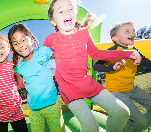 Kinder in der Hüpfburg Vier Kinder springen lachend zusammen auf einer grünen Hüpfburg im Freien bei Sonnenschein.