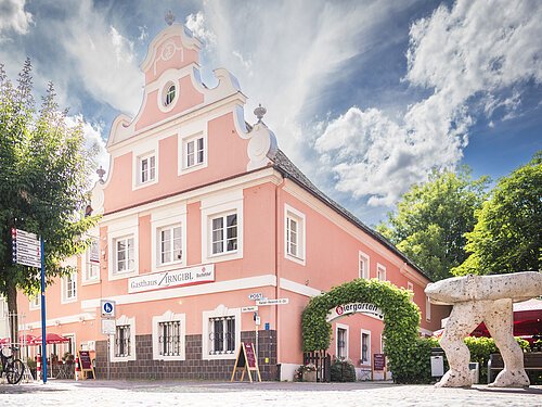 Zirngibl Ein rosa Gebäude mit Barockfassade, daneben ein Biergarten mit grüner Laube, im Vordergrund eine Steinfigur.