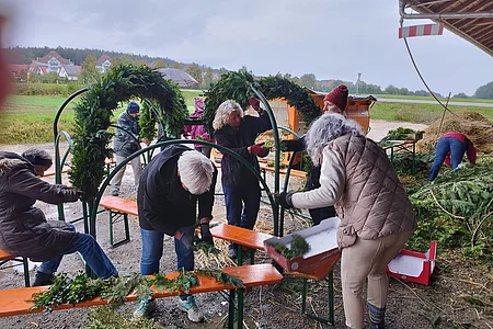 Mehrere Personen binden draußen an Metallrahmen grüne Zweige zu Dekorationen bei bewölktem Himmel.