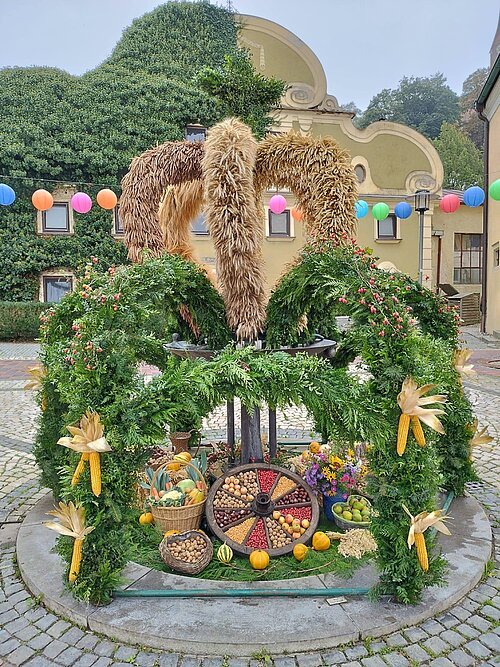 Erntedankkrone am Marktplatz Erntekrone aus Getreide und grünen Zweigen mit Früchten und Gemüse auf gepflastertem Platz vor Hausfassade.
