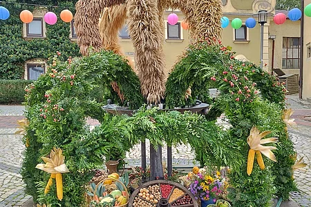 Erntekrone aus Getreide und grünen Zweigen mit Früchten und Gemüse auf gepflastertem Platz vor Hausfassade.