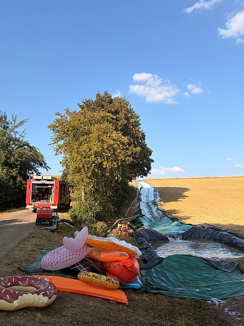 Wasserrutsche in Saalhaupt Wasserrutsche mit Pool, Feuerwehrfahrzeug und Bäumen bei klarem Himmel auf Feldweg im Freien.