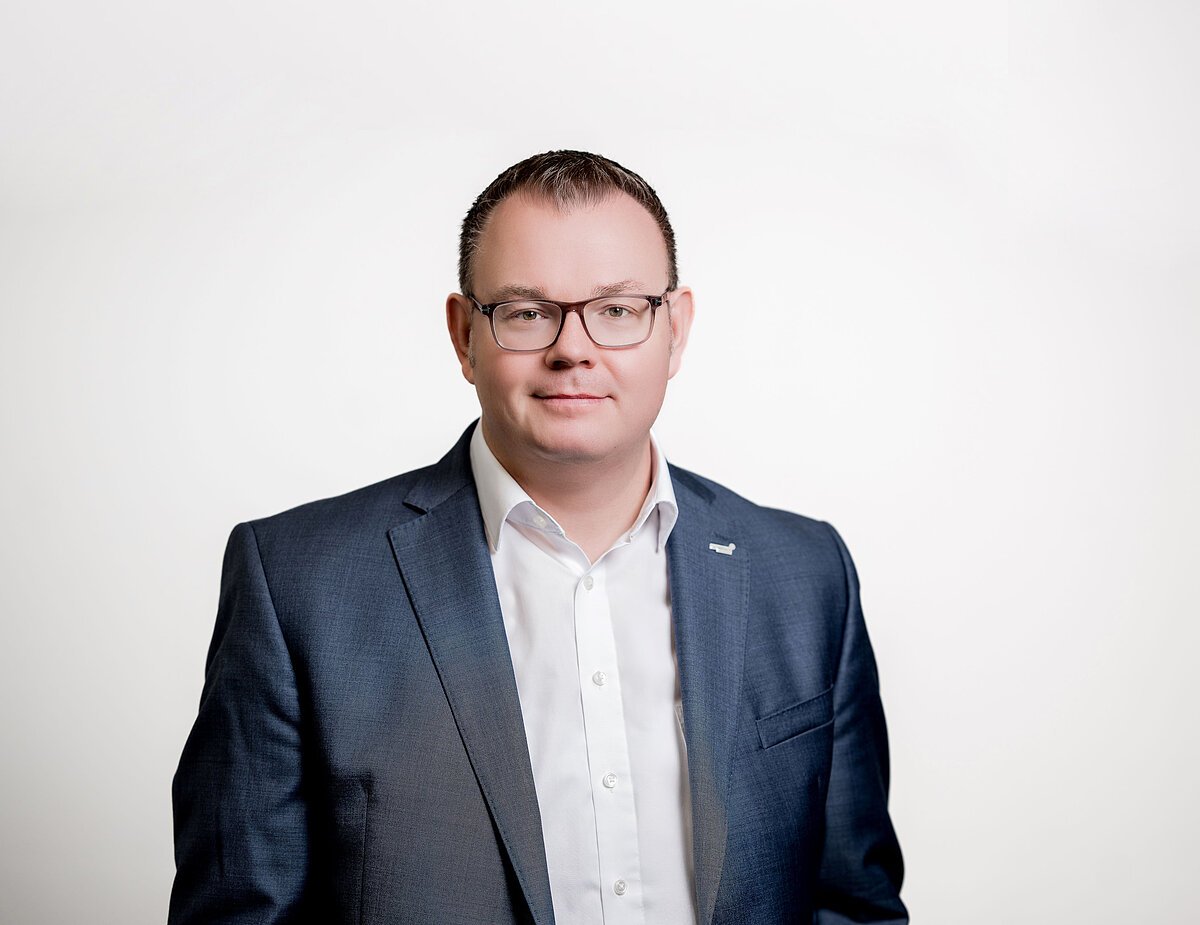 Dr. Benedikt Grünewald Porträt eines Mannes mit Brille, weißem Hemd und blauem Sakko vor weißem Hintergrund.