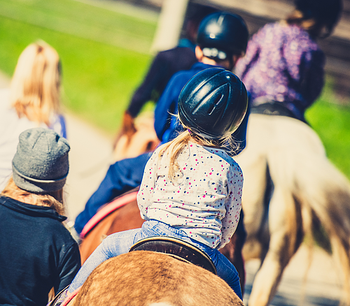 Kinder beim geführten Ausritt Kinder mit Reithelmen reiten hintereinander auf Pferden, eine erwachsene Person begleitet sie zu Fuß.