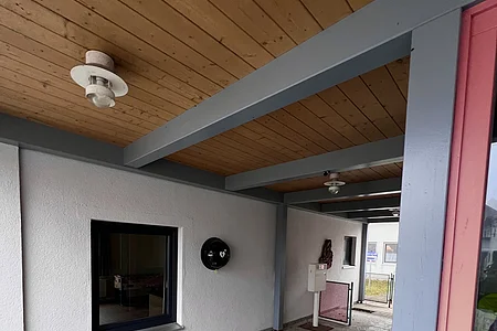 Überdachter Hauseingang mit grauem Boden, weißer Wand, Holzdecke und Briefkasten auf einem Pfosten.
