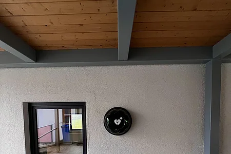Defibrillator in schwarzem Gehäuse an weißer Wand neben Fenster in überdachtem Außenbereich.