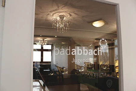 Glasfenster mit Aufschrift „bad abbach Jugendtreff“ und Spiegelung eines Raumes mit Kronleuchtern und Tischkicker