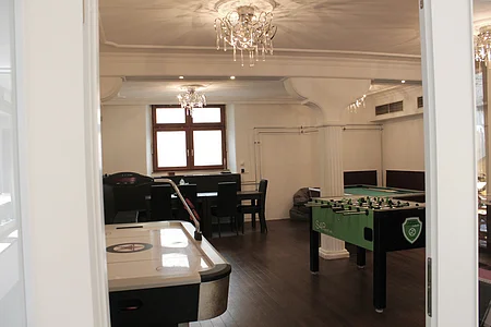 Blick in einen Raum mit Airhockey-Tisch, Kicker, Sitzbank, Tisch und Stühlen vor einem Fenster.
