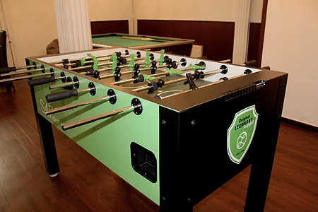 Kicker-Tisch mit grünen und schwarzen Spielfiguren in einem Raum mit Holzboden und Billardtisch im Hintergrund.