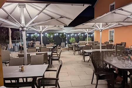 Außenbereich eines Restaurants mit Tischen, Stühlen, großen Sonnenschirmen und Beleuchtung bei Nacht.
