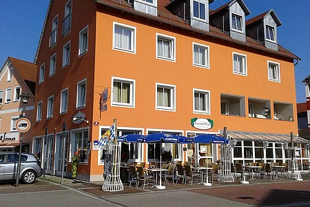 Oranger mehrstöckiger Gebäudekomplex mit Außengastronomie und blauen Sonnenschirmen an sonnigem Tag.