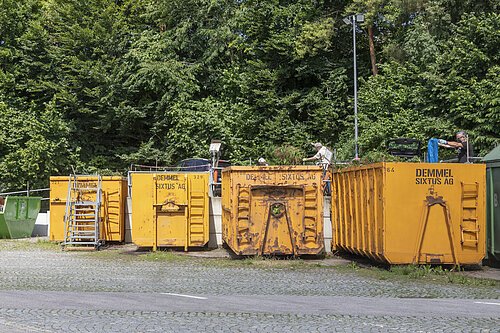 Vier gelbe Container mit der Aufschrift Demmel Sixtus AG stehen vor einem Wald, zwei Personen befüllen sie.