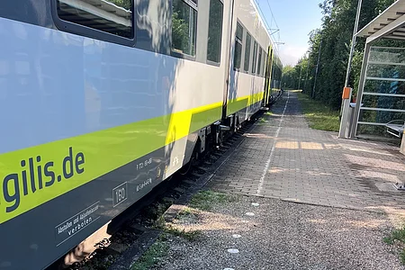Nahaufnahme eines stehenden Zuges mit gelbem Streifen an einem Bahnsteig mit Kiesweg und Wartehäuschen bei Sonnenschein.