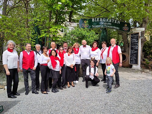 Gospelchor Call It Out Gruppenfoto von 16 Personen vor einem Biergarten-Schild, viele tragen weiße Oberteile mit roten Accessoires, draußen im Grünen.