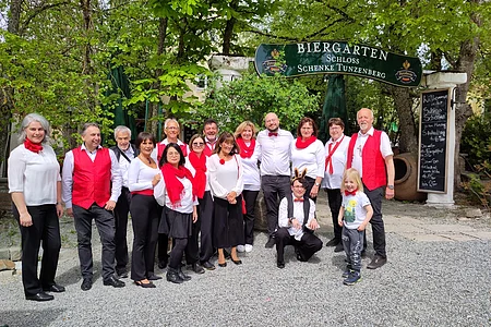 Gruppenfoto von 16 Personen vor einem Biergarten-Schild, viele tragen weiße Oberteile mit roten Accessoires, draußen im Grünen.