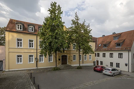 Gelbes zweistöckiges Gebäude mit braunem Dach, zwei Bäumen davor und zwei geparkten Autos rechts.