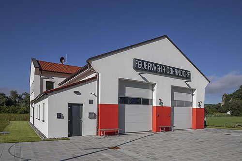 Freiwillige Feuerwehr Oberndorf e. V. Feuerwehrgebäude Oberndorf mit zwei großen Toren und rotem Streifen an der Fassade bei klarem Himmel.