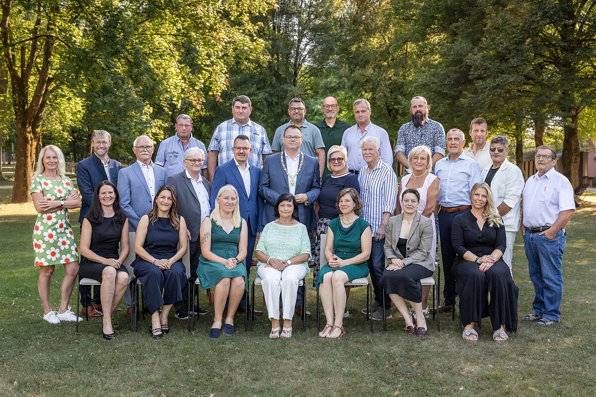 Gruppenfoto von 29 Personen, Männer und Frauen, im Freien vor Bäumen, einige sitzen auf Stühlen, andere stehen.