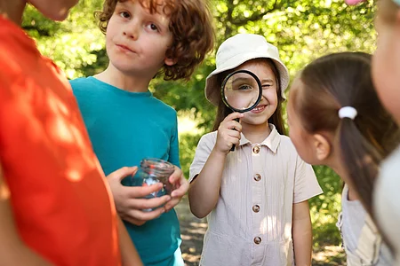 Kinder im Freien, ein Mädchen schaut durch eine Lupe, ein Junge hält ein Glas, andere Kinder stehen um sie herum.