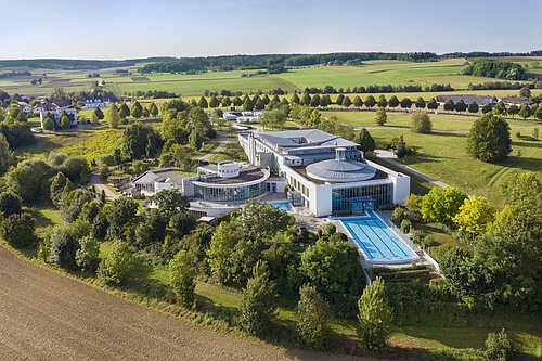 Luftaufnahme Kaiser-Therme Luftaufnahme eines modernen Gebäudekomplexes mit Außenpool umgeben von Bäumen und Feldern.