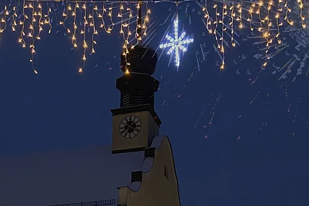 Kirchturm mit Uhr bei Nacht, darüber Lichterketten und ein leuchtender Stern, im Hintergrund Feuerwerk