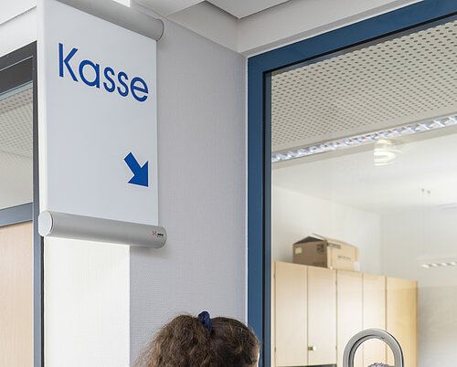 Frau an Kasse mit Formular, spricht mit Mitarbeiterin hinter Glasscheibe in Büro mit Regalen und Kartons.
