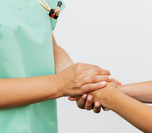 Hände eines medizinischen Fachpersonals in grüner Kleidung halten ein Patientenhandgelenk mit weißem Hintergrund.